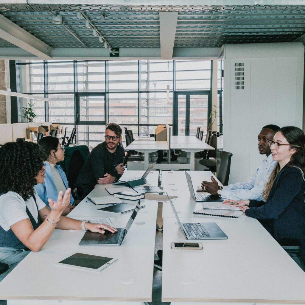 Un equipo diverso de cinco profesionales sentados en una mesa de oficina abierta, discutiendo proyectos con laptops y tablets en un ambiente de trabajo colaborativo y moderno.