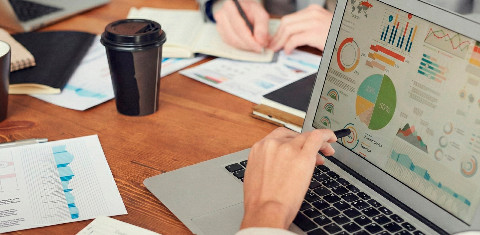analisis-kpis-dashboard-metricas-rendimiento-innk Primer plano de un profesional señalando con un bolígrafo un gráfico circular en la pantalla de una laptop que muestra un dashboard interactivo, con reportes impresos y una taza de café sobre un escritorio de madera.