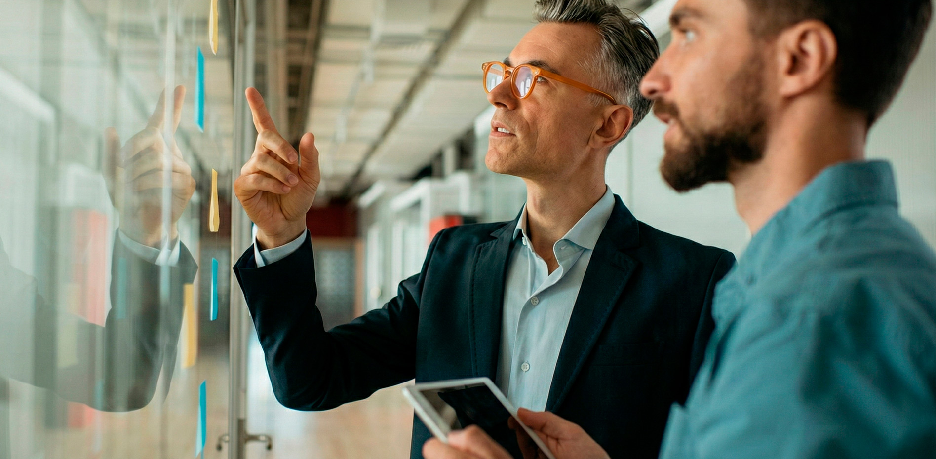 mentoring-estrategico-planificacion-agile-innk Un ejecutivo senior con gafas naranja señala un punto específico en un panel de cristal con notas adhesivas mientras conversa con un colega que sostiene una tablet en una oficina moderna.