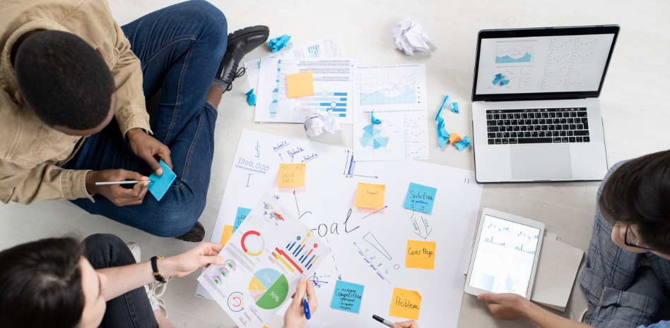Vista aérea de un equipo creativo realizando una sesión de lluvia de ideas en el suelo, rodeados de gráficos impresos, notas adhesivas de colores, una tablet y una laptop, enfocados en definir metas y soluciones estratégicas.