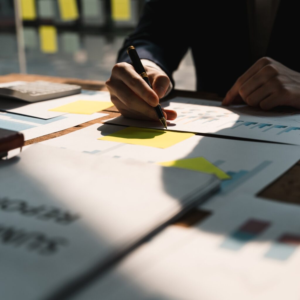 Primer plano de un profesional utilizando una pluma para revisar gráficos de barras y reportes financieros sobre un escritorio de madera, con notas adhesivas amarillas para resaltar datos clave.