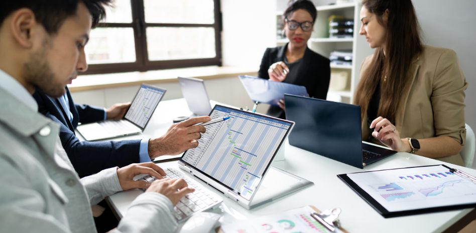 Un equipo de cuatro profesionales colaborando en una mesa de oficina con laptops; en primer plano, se observa un diagrama de Gantt detallado en una pantalla mientras un integrante señala el cronograma de actividades.