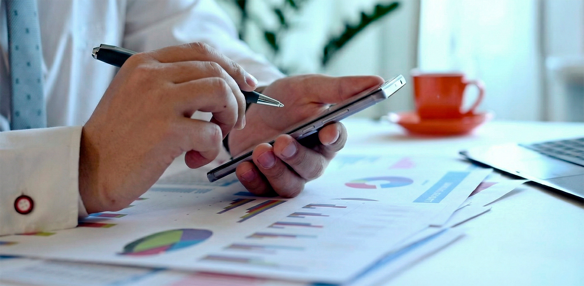 analisis-datos-metricas-smartphone-gestion-proyectos-innovacion Primer plano de un profesional utilizando un smartphone y un bolígrafo para revisar reportes impresos con gráficos de rendimiento y métricas sobre un escritorio, representando el seguimiento analítico y la toma de decisiones en la gestión de proyectos.