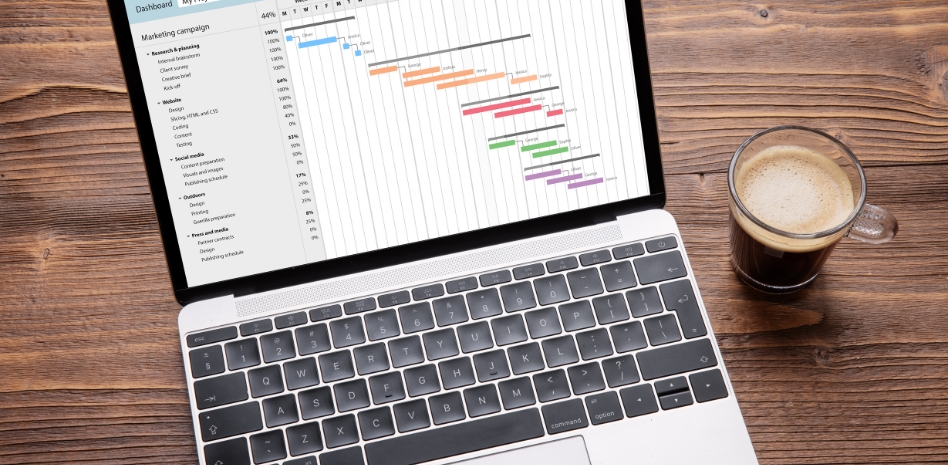 Vista superior de una laptop mostrando un diagrama de Gantt para la planificación detallada de una campaña de marketing, junto a una taza de café sobre un escritorio de madera, representando la productividad y el enfoque en la gestión de proyectos.