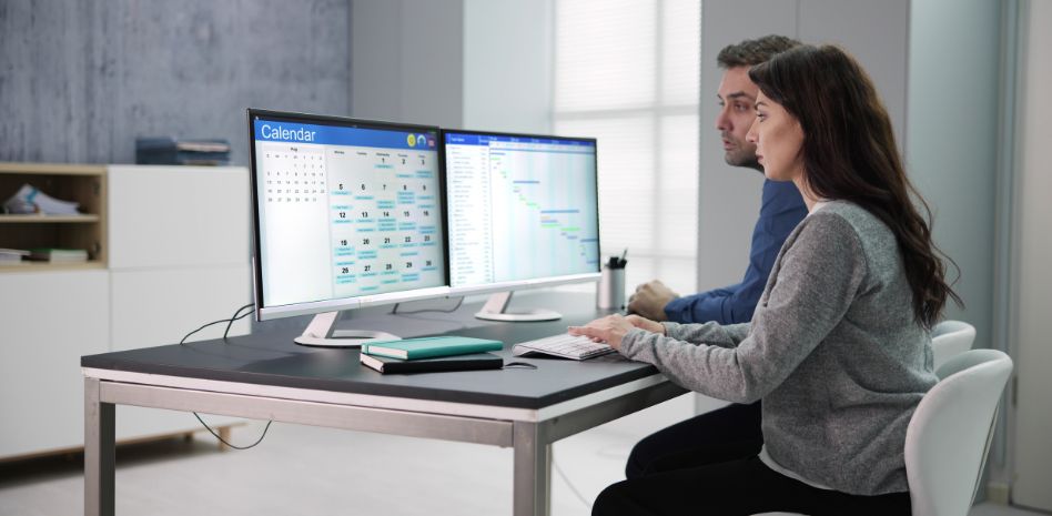 Dos profesionales colaborando en un escritorio con monitores duales, alineando un calendario de actividades con un diagrama de Gantt para la planificación y gestión de proyectos de innovación empresarial.