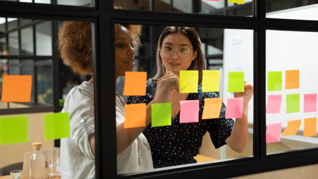 Dos profesionales colaborando en un panel de vidrio con notas adhesivas para la gestión de proyectos, visualizando ideas para impulsar la innovación empresarial y estructurar el portafolio estratégico.