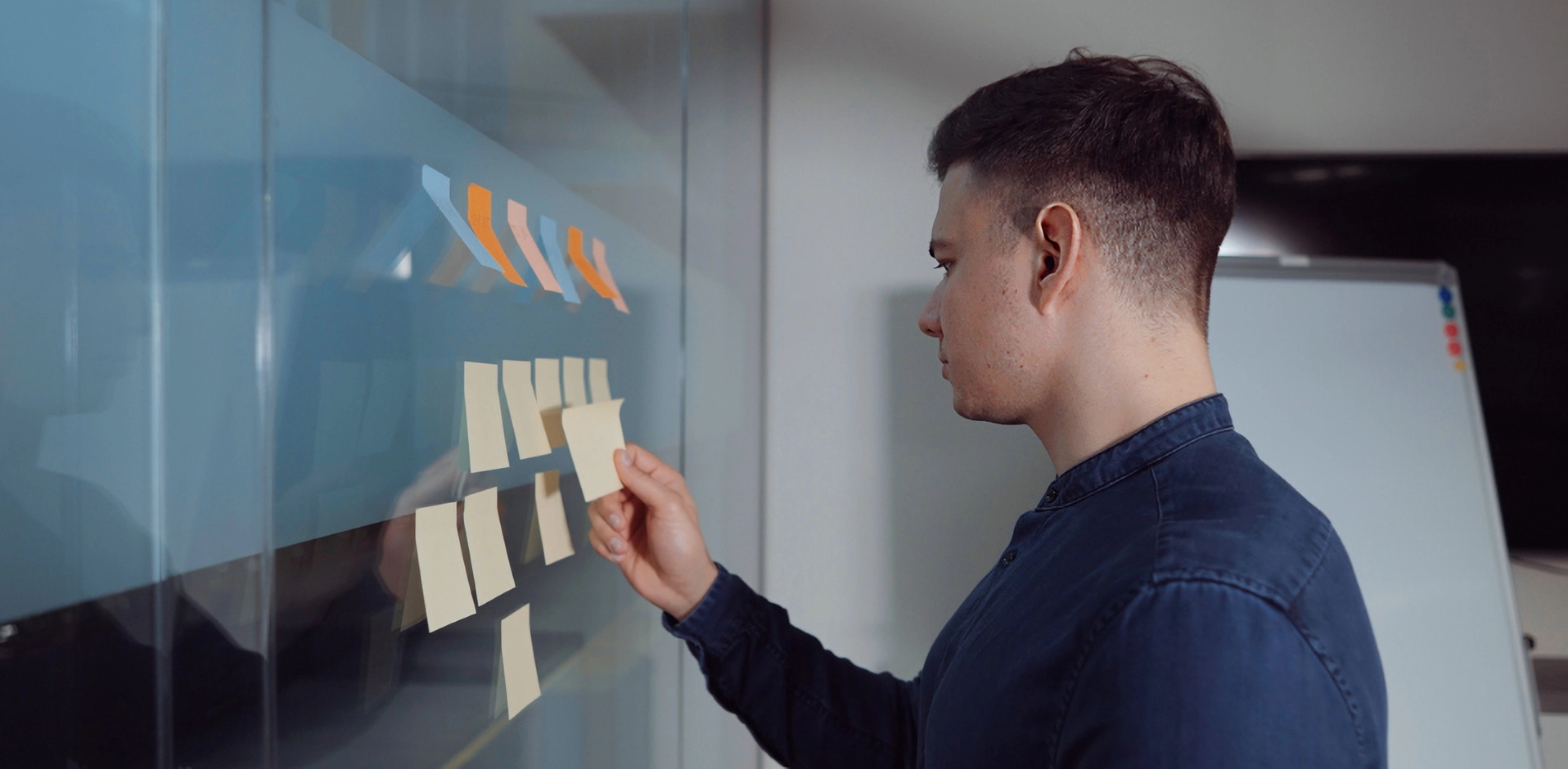 Profesional organizando tareas e ideas con notas adhesivas en una pared de cristal, aplicando metodologías ágiles para la planificación y gestión de proyectos de innovación empresarial.