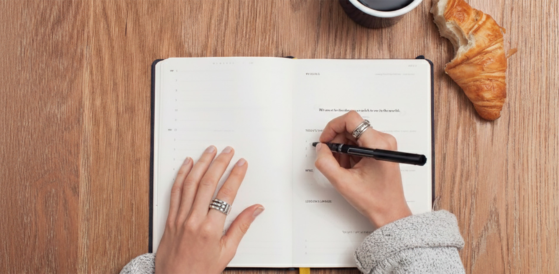 Vista cenital de una persona escribiendo en una agenda abierta sobre una mesa de madera, acompañada de un café y un croissant, representando la planificación diaria y el equilibrio vida-trabajo.