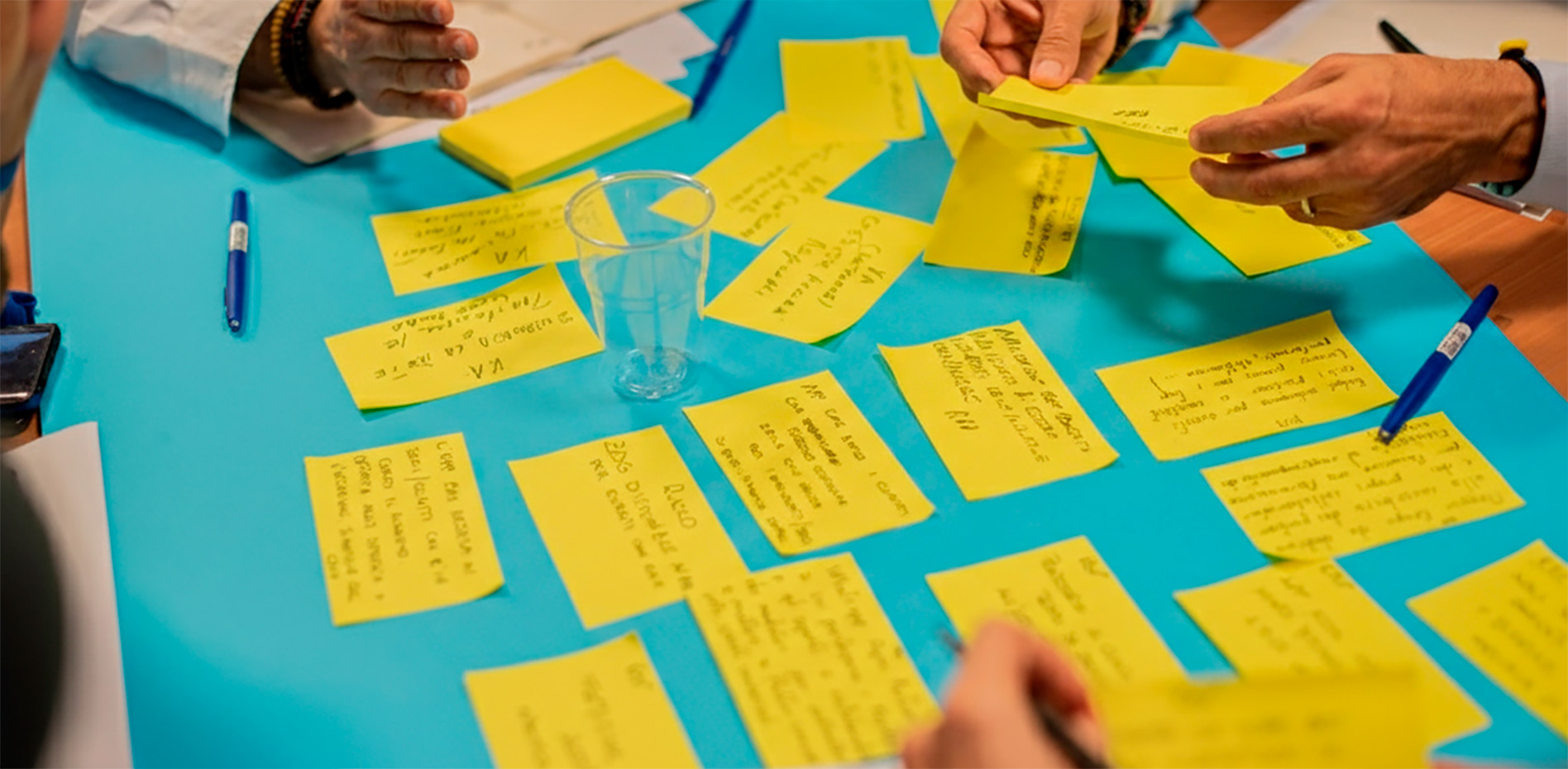 sesion-colaborativa-mapeo-ideas-post-its-innk Vista superior de una mesa azul donde manos de varios profesionales organizan y analizan numerosas notas adhesivas amarillas con anotaciones durante un taller de co-creación.