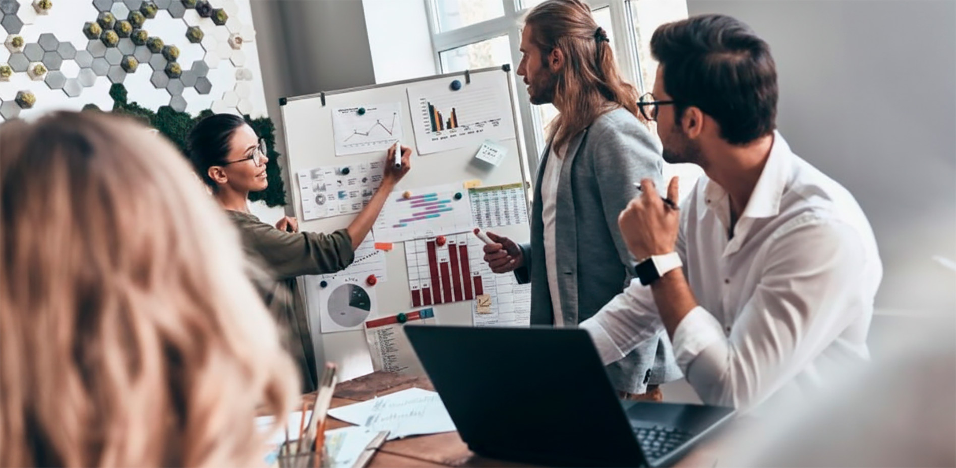 Una profesional explica gráficos de rendimiento y KPIs en una pizarra blanca durante una reunión de equipo en una oficina moderna.