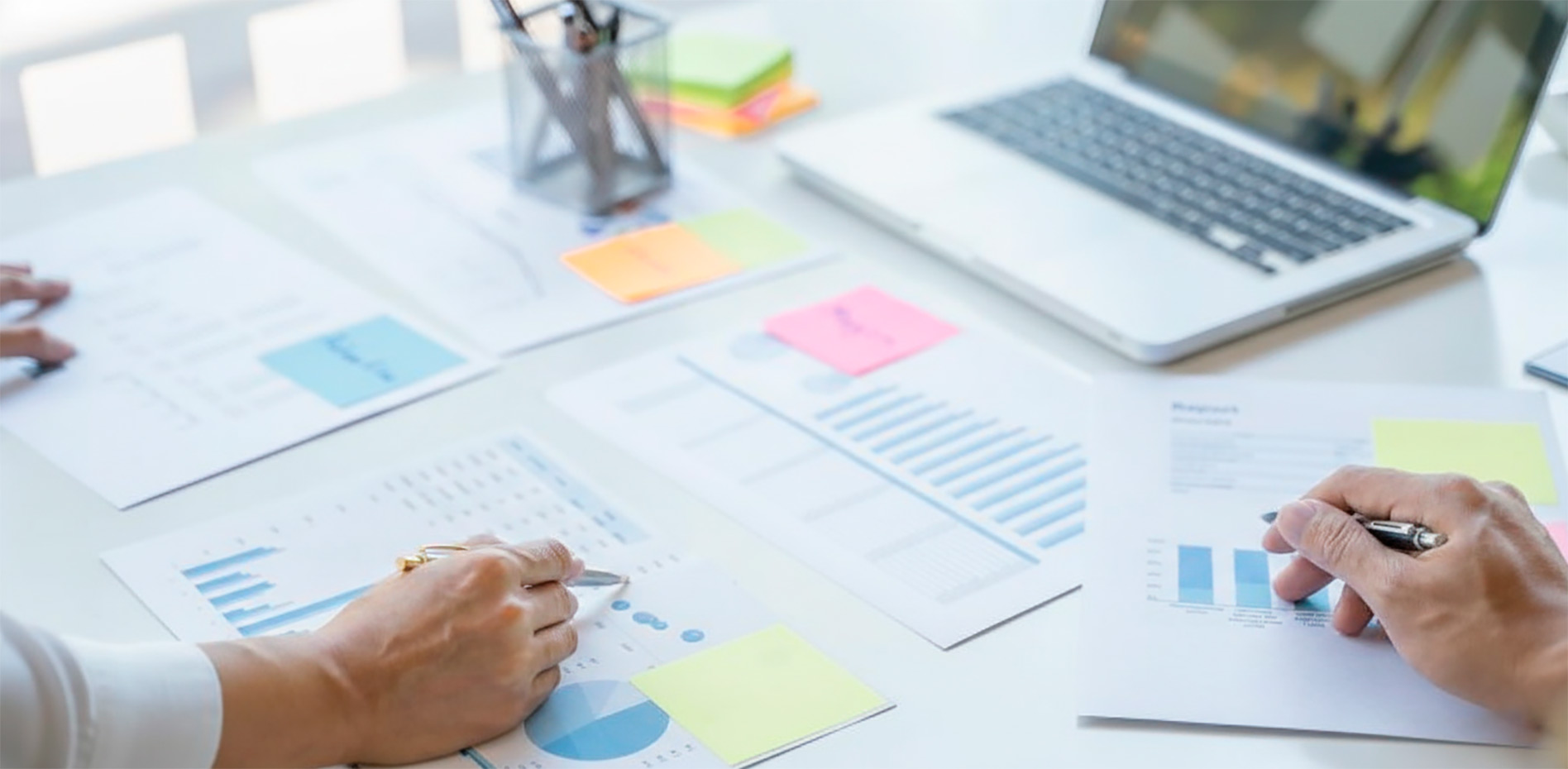 analisis-reportes-financieros-gestion-proyectos-innk Primer plano de manos de profesionales analizando reportes con gráficos de barras, diagramas circulares y notas adhesivas sobre una mesa de trabajo con laptop.