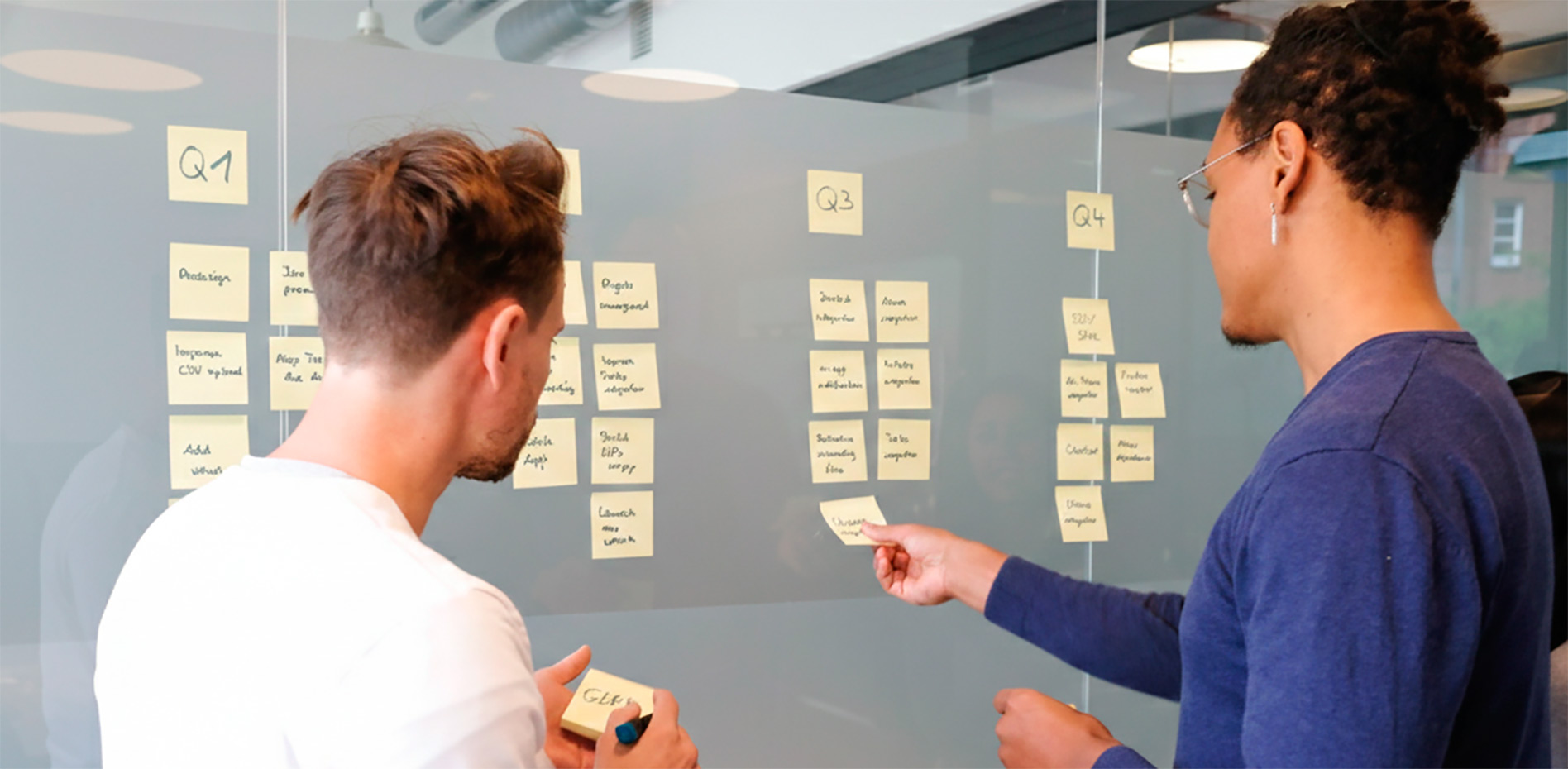 planificacion-trimestral-roadmap-equipo-proyectos-innk Dos profesionales organizan tareas en un panel de cristal dividido por trimestres (Q1 a Q4) utilizando notas adhesivas amarillas en una oficina moderna.