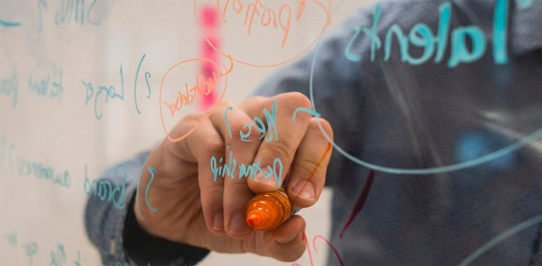 procesos-mentoria-talento-estrategia-negocios-innk Primer plano de una mano escribiendo conceptos de talento, mentoria y alianzas estratégicas con un rotulador naranja sobre una pizarra de cristal transparente.