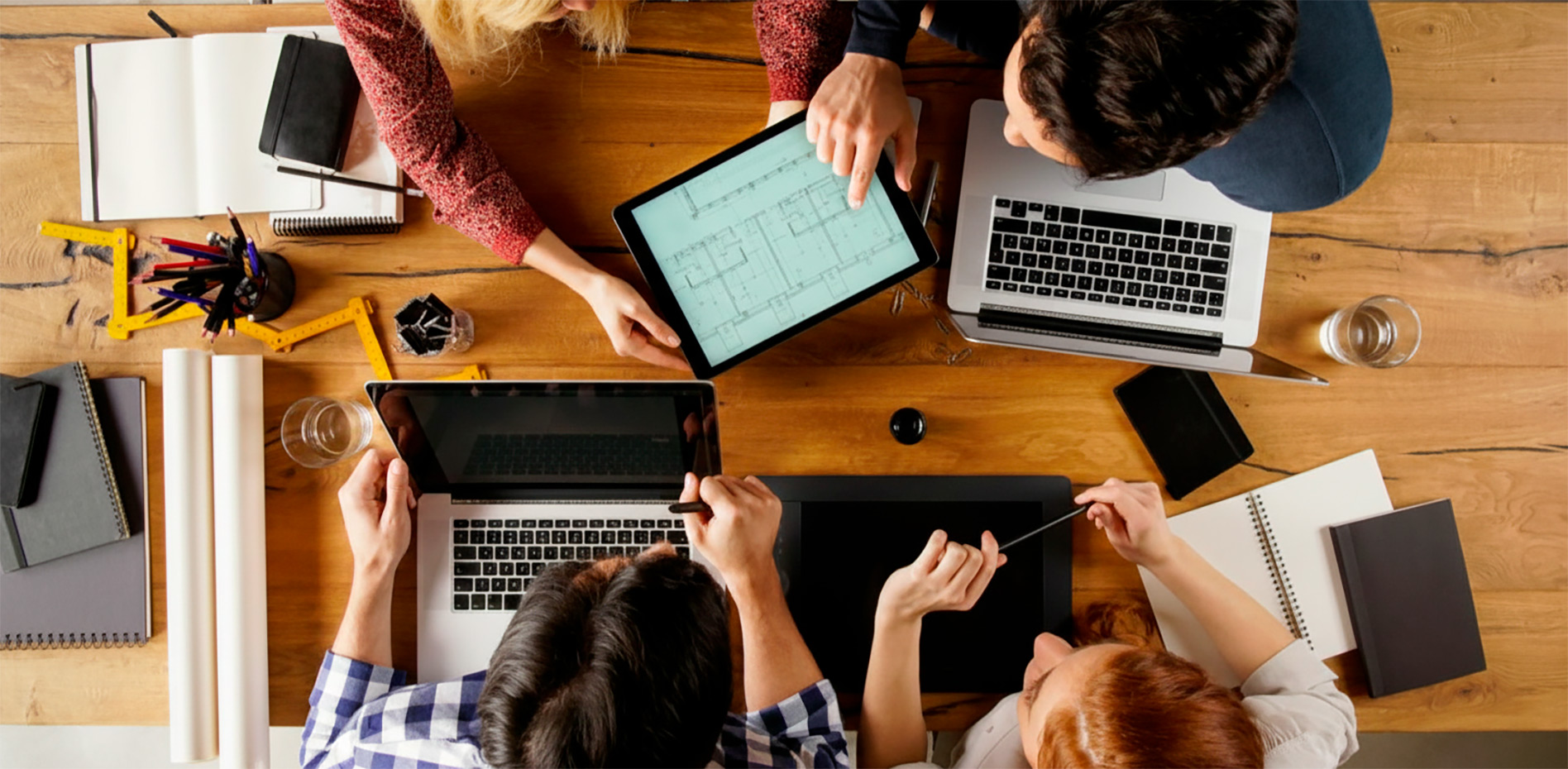 Vista aérea de un equipo de cuatro personas trabajando colaborativamente en una mesa de madera con laptops, una tablet mostrando un plano técnico, cuadernos y herramientas de dibujo.