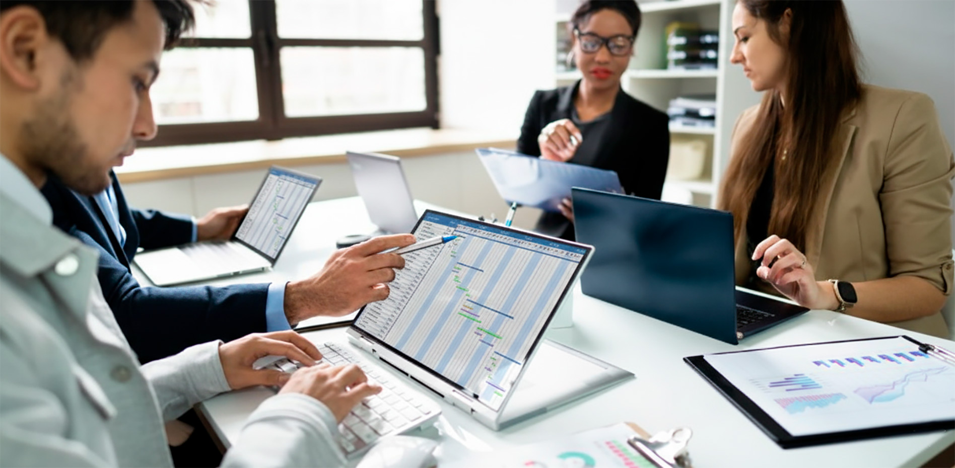 equipo-trabajo-colaborativo-planificacion-proyectos-innk Un grupo de profesionales en una reunión de trabajo colaborativo; un hombre señala un diagrama de Gantt en una laptop convertible mientras otros colegas operan sus computadoras y revisan reportes impresos con gráficos.