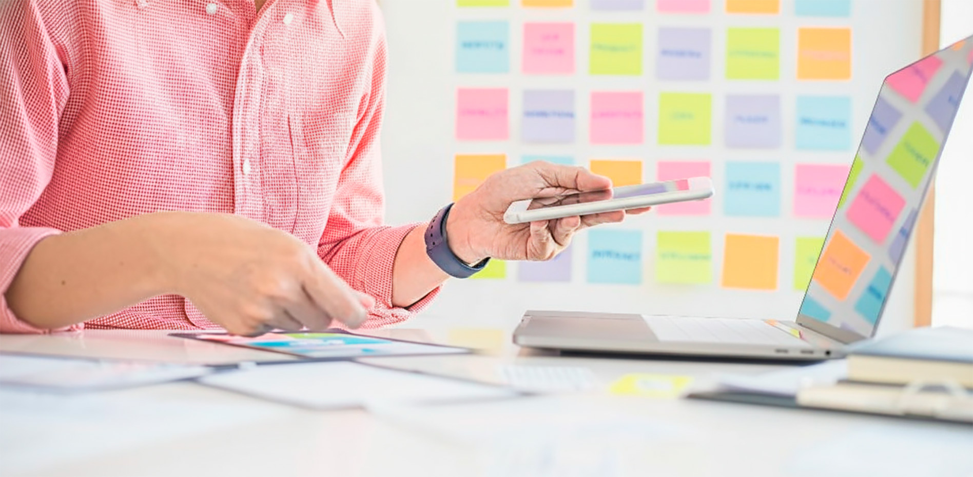 integracion-dispositivos-moviles-gestion-agile-innk Un profesional utiliza un smartphone mientras trabaja frente a una laptop en un escritorio, con una pizarra de notas adhesivas de colores al fondo para organización de tareas.