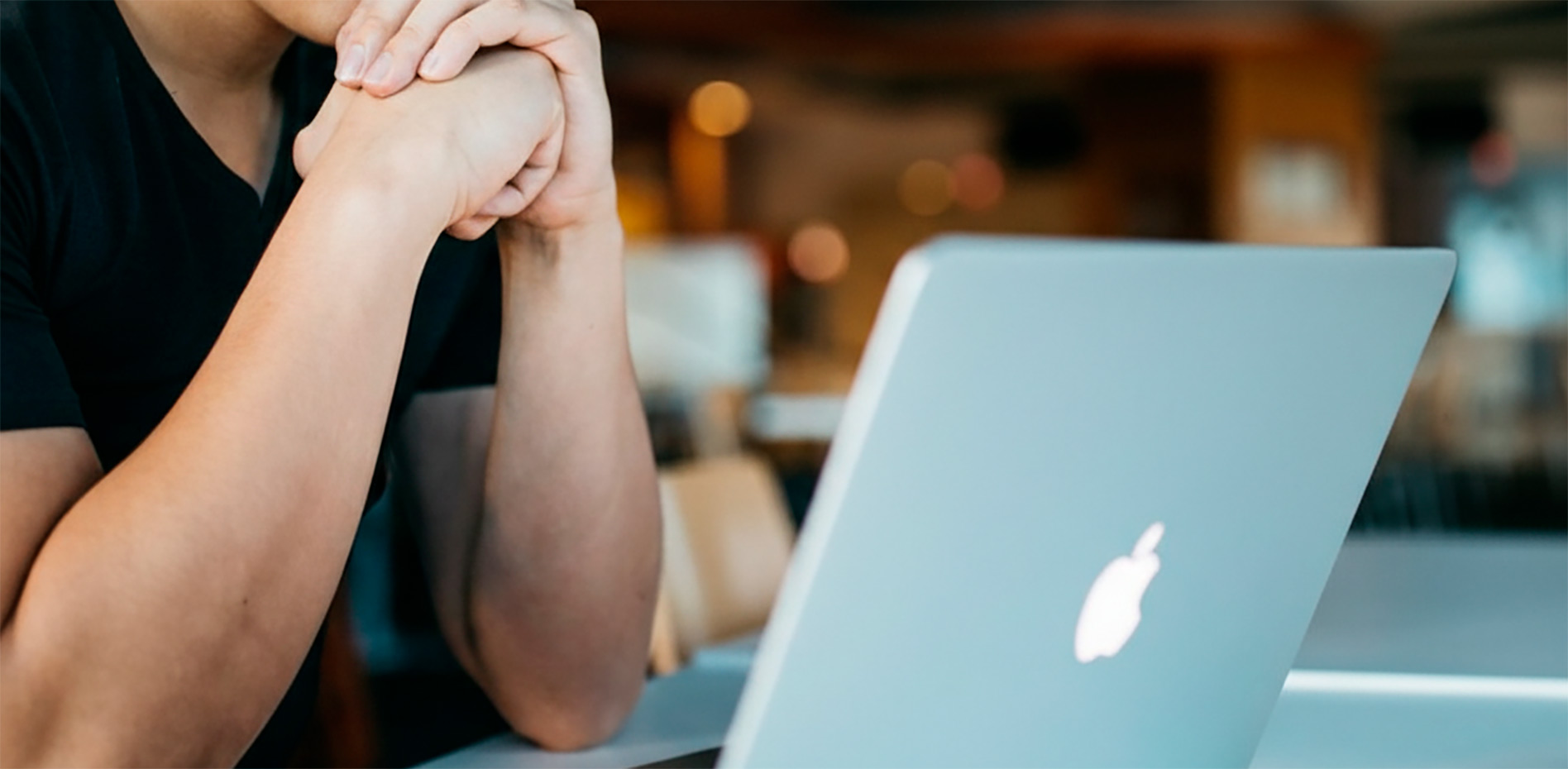 concentracion-trabajo-remoto-laptop-oficina-innk Primer plano de los brazos y manos entrelazadas de un profesional sentado frente a una laptop abierta en un ambiente de oficina o café con fondo desenfocado.