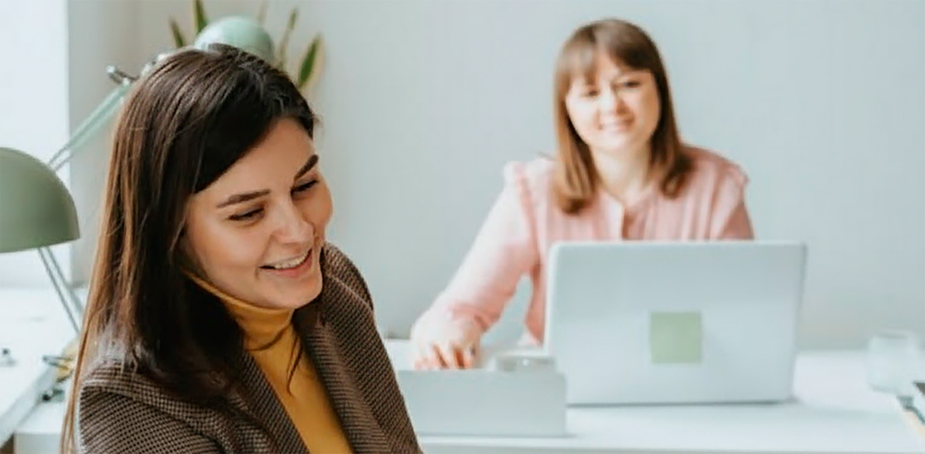 colaboracion-equipo-trabajo-hibrido-oficina-innk Dos profesionales colaboran en una oficina moderna; una mujer sonríe en primer plano mientras su colega trabaja con una laptop en un escritorio luminoso al fondo.